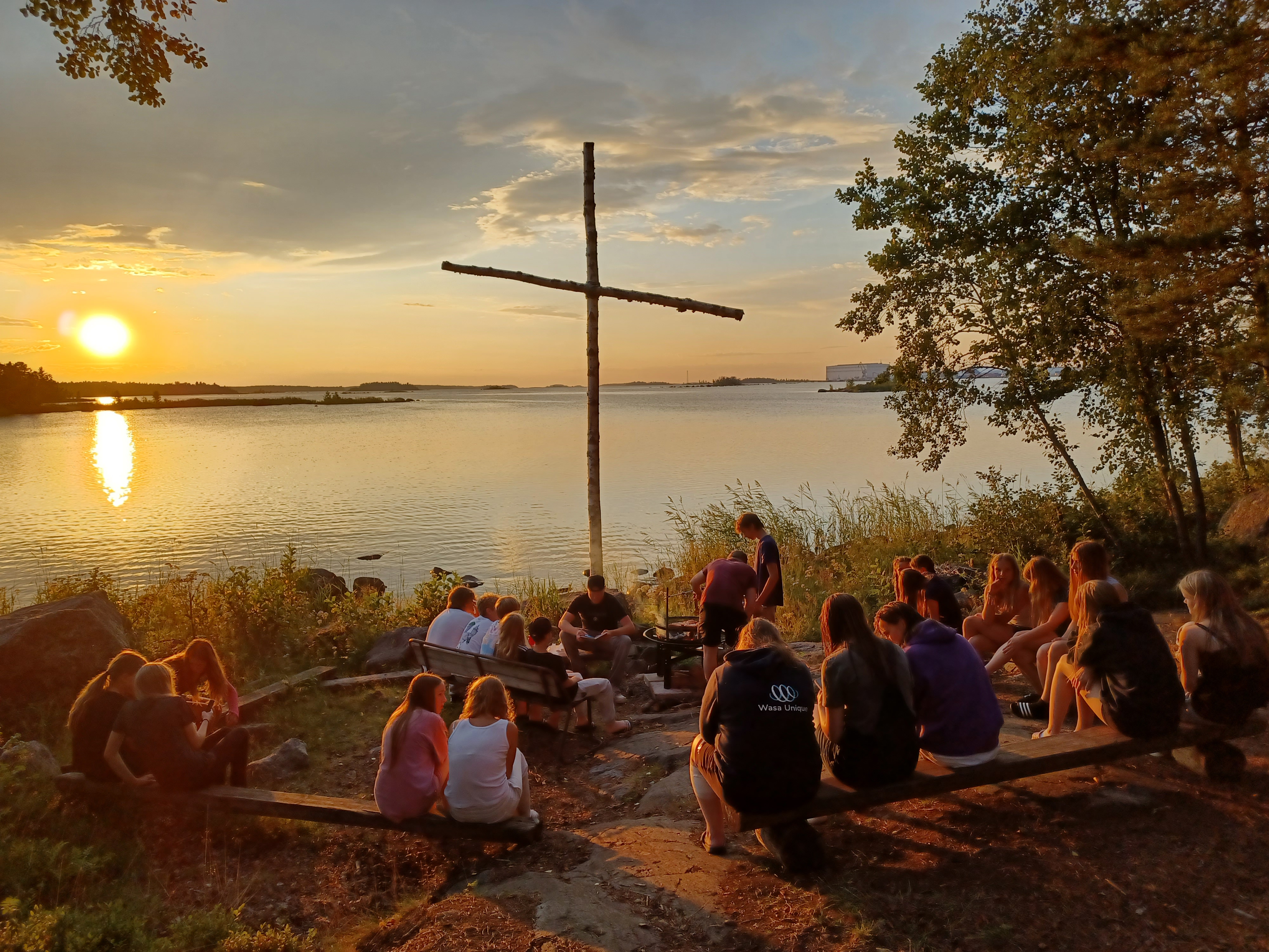 Lägerdeltagare vid kvällsbålet vid sjöstranden. I bakgrunden ett träkors.