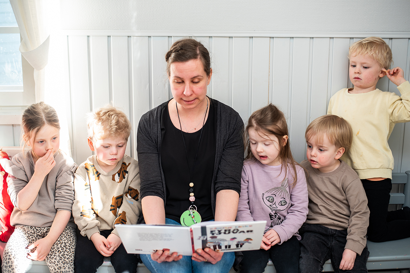 Lärare Ingela  Silfverberg sitter på en bänk och läser en bok för fem barn som sitter på var sida om henne.